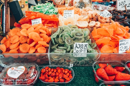 A vibrant market stall displaying an assortment of dried fruits, such as apricots, kiwis, and mangos. Fresh strawberries are also visible in plastic containers. Each section has price tags indicating the cost of the items.