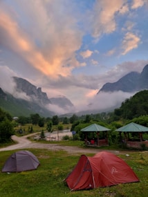 A scenic landscape with a lush green valley surrounded by towering mountains partially shrouded in mist. The sky displays a blend of soft pastel hues with clouds illuminated by the setting or rising sun. In the foreground, there are two tents set up in a grassy area near wooden gazebos with green roofs, adjacent to a dirt path.