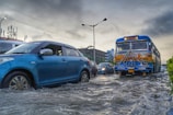 Several vehicles are driving through a flooded street. A blue car is prominently visible on the left side, while a decorated bus is to the right. The water level is quite high, submerging the tires of the vehicles. The sky is cloudy and dark, indicating a recent or ongoing storm. Buildings and street lamps are visible in the background.