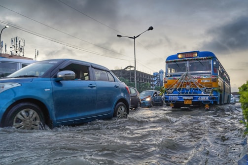 Several vehicles are driving through a flooded street. A blue car is prominently visible on the left side, while a decorated bus is to the right. The water level is quite high, submerging the tires of the vehicles. The sky is cloudy and dark, indicating a recent or ongoing storm. Buildings and street lamps are visible in the background.