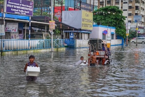 A flooded street scene in an urban area, with several children wading through knee-deep water. One child carries a Styrofoam box, while another group of children is gathered around a cycle rickshaw. Buildings and street signs are visible in the background, indicating a busy and populated area.
