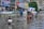 A flooded street scene in an urban area, with several children wading through knee-deep water. One child carries a Styrofoam box, while another group of children is gathered around a cycle rickshaw. Buildings and street signs are visible in the background, indicating a busy and populated area.