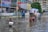 A flooded street scene in an urban area, with several children wading through knee-deep water. One child carries a Styrofoam box, while another group of children is gathered around a cycle rickshaw. Buildings and street signs are visible in the background, indicating a busy and populated area.
