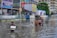 A flooded street scene in an urban area, with several children wading through knee-deep water. One child carries a Styrofoam box, while another group of children is gathered around a cycle rickshaw. Buildings and street signs are visible in the background, indicating a busy and populated area.