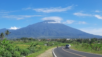 A scenic mountain landscape with a clear blue sky, a winding road leading towards the mountain, and lush green fields on either side. A few vehicles and motorbikes travel along the road, with the mountain dominating the background. Light clouds hover around the mountain peak, adding to the serene atmosphere.
