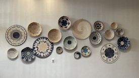 A collection of decorative, woven baskets with various intricate patterns and designs arranged on a light-colored wall. The patterns feature geometric and symmetrical motifs, primarily in black, beige, and natural hues.