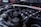 A close-up view of a car engine, showing various components including the intake manifold, hoses, and a brace bar with a hashtag. The background features parts of a car, likely in a garage or outdoor setting.