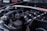 A close-up view of a car engine, showing various components including the intake manifold, hoses, and a brace bar with a hashtag. The background features parts of a car, likely in a garage or outdoor setting.