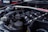 A close-up view of a car engine, showing various components including the intake manifold, hoses, and a brace bar with a hashtag. The background features parts of a car, likely in a garage or outdoor setting.