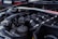 A close-up view of a car engine, showing various components including the intake manifold, hoses, and a brace bar with a hashtag. The background features parts of a car, likely in a garage or outdoor setting.