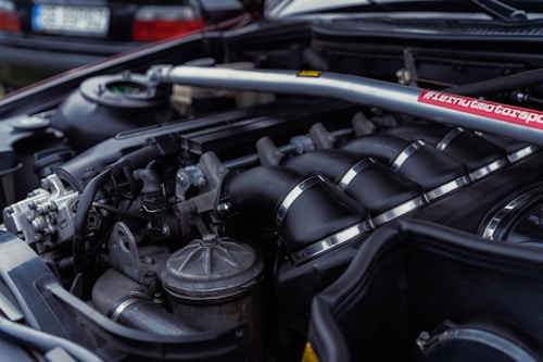 A close-up view of a car engine, showing various components including the intake manifold, hoses, and a brace bar with a hashtag. The background features parts of a car, likely in a garage or outdoor setting.