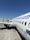 A white commercial airplane is parked on the tarmac at an airport. The fuselage features a website URL and flag graphics near the tail. The airplane is positioned parallel to the terminal building, showing its wing and landing gear. The sky is clear and blue, providing a bright and sunny atmosphere.