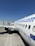 A white commercial airplane is parked on the tarmac at an airport. The fuselage features a website URL and flag graphics near the tail. The airplane is positioned parallel to the terminal building, showing its wing and landing gear. The sky is clear and blue, providing a bright and sunny atmosphere.