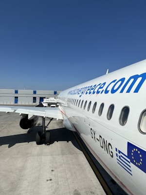A white commercial airplane is parked on the tarmac at an airport. The fuselage features a website URL and flag graphics near the tail. The airplane is positioned parallel to the terminal building, showing its wing and landing gear. The sky is clear and blue, providing a bright and sunny atmosphere.