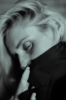 A person with closed eyes rests their head on a dark fabric, with fingers gently touching the material. The lighting creates a soft, intimate mood.
