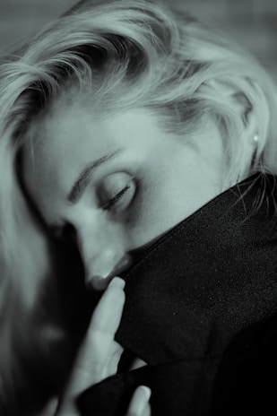 A person with closed eyes rests their head on a dark fabric, with fingers gently touching the material. The lighting creates a soft, intimate mood.