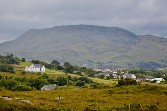A peaceful rural landscape featuring a cluster of houses scattered across rolling green hills. A prominent white house stands out among the greenery, with a backdrop of towering mountains under a cloudy sky.