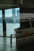 A reflective glass surface displays a layered image with an indoor scene blending into an outdoor view. Inside, a van with a colorful stripe is parked and partially visible with a stool and a small table holding drinks. Outside, a city skyline can be faintly seen in the background, with prominent tall buildings.