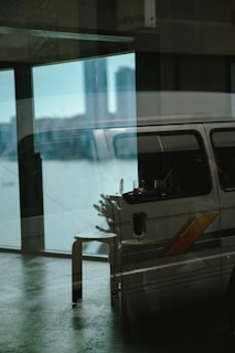 A reflective glass surface displays a layered image with an indoor scene blending into an outdoor view. Inside, a van with a colorful stripe is parked and partially visible with a stool and a small table holding drinks. Outside, a city skyline can be faintly seen in the background, with prominent tall buildings.