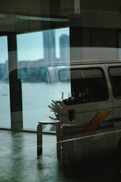 A reflective glass surface displays a layered image with an indoor scene blending into an outdoor view. Inside, a van with a colorful stripe is parked and partially visible with a stool and a small table holding drinks. Outside, a city skyline can be faintly seen in the background, with prominent tall buildings.