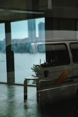 A reflective glass surface displays a layered image with an indoor scene blending into an outdoor view. Inside, a van with a colorful stripe is parked and partially visible with a stool and a small table holding drinks. Outside, a city skyline can be faintly seen in the background, with prominent tall buildings.