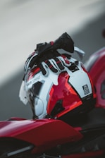A red and white motorcycle helmet rests on a motorcycle, with a black and red glove placed on top. The helmet has a sleek design and the setting appears to be outdoors on a paved surface.