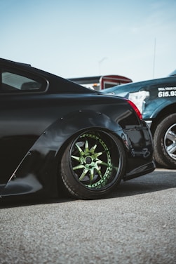 A low-riding black car with green custom wheels is prominently featured. It is parked on a paved surface next to another vehicle. The car has a sleek, glossy finish and a modified fender. The background includes part of another car and some outdoor elements.