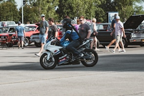 A person wearing a helmet and motorcycle gear rides a white sport bike labeled with logos in an open parking lot. In the background, several classic cars are on display with their hoods open. People are walking around, examining the vehicles and conversing with each other. The setting appears to be an automotive event or car show.