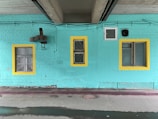 A turquoise-painted brick wall features three windows with yellow frames. A large exhaust fan or ventilation unit is mounted to the wall. There is a section of concrete ceiling above with visible pipes and wiring.