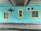 A turquoise-painted brick wall features three windows with yellow frames. A large exhaust fan or ventilation unit is mounted to the wall. There is a section of concrete ceiling above with visible pipes and wiring.