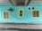A turquoise-painted brick wall features three windows with yellow frames. A large exhaust fan or ventilation unit is mounted to the wall. There is a section of concrete ceiling above with visible pipes and wiring.