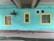 A turquoise-painted brick wall features three windows with yellow frames. A large exhaust fan or ventilation unit is mounted to the wall. There is a section of concrete ceiling above with visible pipes and wiring.