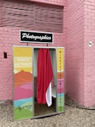 A vintage-style photo booth is situated against a pink brick wall. The booth features colorful panels with abstract art patterns and a curtain that is red and white. It has a retro sign on top reading 'Photographies.'