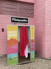 A vintage-style photo booth is situated against a pink brick wall. The booth features colorful panels with abstract art patterns and a curtain that is red and white. It has a retro sign on top reading 'Photographies.'