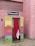 A vintage-style photo booth is situated against a pink brick wall. The booth features colorful panels with abstract art patterns and a curtain that is red and white. It has a retro sign on top reading 'Photographies.'
