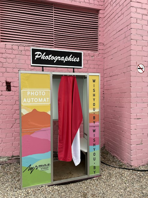 A vintage-style photo booth is situated against a pink brick wall. The booth features colorful panels with abstract art patterns and a curtain that is red and white. It has a retro sign on top reading 'Photographies.'