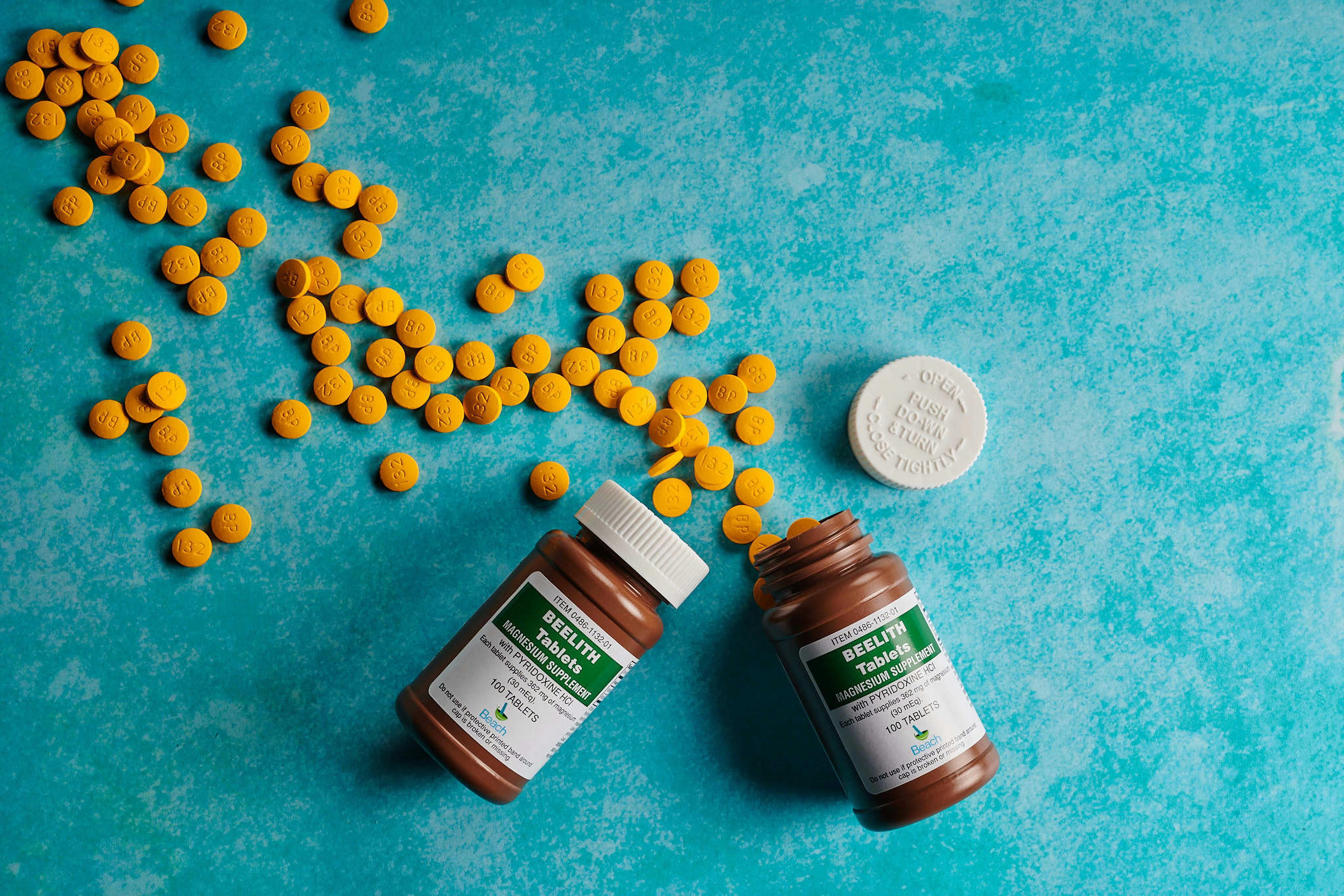 Several yellow pills are scattered across a textured, turquoise background. Two brown pill bottles with green labels are open, surrounded by the pills. A white bottle cap is visible next to the bottles.