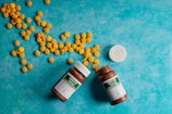 Several yellow pills are scattered across a textured, turquoise background. Two brown pill bottles with green labels are open, surrounded by the pills. A white bottle cap is visible next to the bottles.