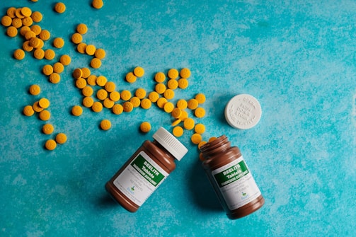 Several yellow pills are scattered across a textured, turquoise background. Two brown pill bottles with green labels are open, surrounded by the pills. A white bottle cap is visible next to the bottles.