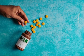 A hand is reaching for a scattered group of yellow tablets on a textured turquoise surface. A brown bottle labeled 'Beelith Tablets' is lying on its side, partly empty.
