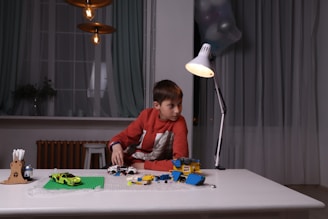 A young boy with brown hair is seated at a table, engaged in playing with colorful toy blocks and miniature cars. The room is softly lit by a modern desk lamp, with curtains partially drawn over large windows in the background. Overhead, three pendant lights hang. The setting conveys a cozy and focused atmosphere.