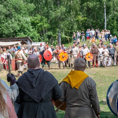 A group of people dressed in historical clothing gathered in an outdoor setting. Some individuals in the front have their backs to the camera, while the majority in the background appear to be engaged in a reenactment or event. Several people are holding shields and wear helmets, and there is lush green vegetation in the background.