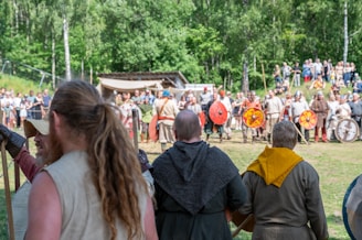 A group of people dressed in historical medieval attire are participating in a reenactment event. The people in the foreground are watching or preparing to join the demonstration, while those in the background seem to be engaged in some form of organized activity, such as a mock battle. Trees and spectators create a natural and lively setting.