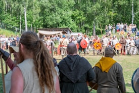 A group of people dressed in historical medieval attire are participating in a reenactment event. The people in the foreground are watching or preparing to join the demonstration, while those in the background seem to be engaged in some form of organized activity, such as a mock battle. Trees and spectators create a natural and lively setting.