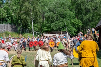 A large group of people are engaged in a historical reenactment scene, with participants dressed in medieval-style clothing and armor, including colorful tunics and cloaks. Many hold round shields, some of which are brightly painted with red and black designs. The event takes place in an open, grassy area surrounded by trees, with onlookers and participants in the background.