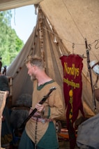 A person dressed in historical or medieval-style clothing, holding a long black object that resembles a staff or weapon. The setting appears to be a canvas tent, possibly part of a historical reenactment or medieval fair. A red banner with the word 'Nordlys' and a website address is visible in the background, alongside various items like furs and medieval props.