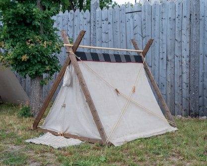 A small, simple canvas tent is set up on a grassy area near a sturdy wooden fence. The tent has a triangular shape with wooden poles providing structure. A green leafy tree stands next to the tent, and there is a mat placed at the entrance.