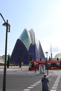 An architectural structure with a modern design featuring a large, triangular building with a combination of glass and blue surfaces. A red tram labeled 'metrovalencia' is moving along the tracks in the foreground. A woman is crossing a street, pushing a shopping trolley, and several tall, slender trees are scattered around.
