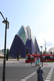 An architectural structure with a modern design featuring a large, triangular building with a combination of glass and blue surfaces. A red tram labeled 'metrovalencia' is moving along the tracks in the foreground. A woman is crossing a street, pushing a shopping trolley, and several tall, slender trees are scattered around.