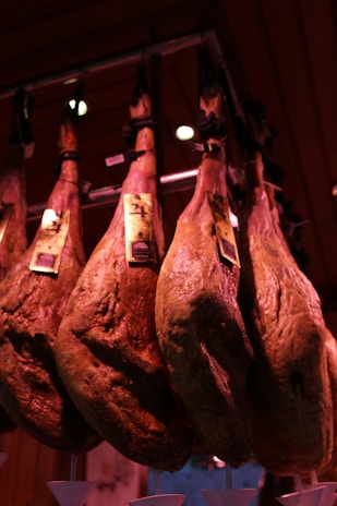 Several large cured hams hang from hooks in a dimly lit room. Each ham has a label attached near the top, and the lighting casts a warm, reddish glow over the scene. They appear to be part of a traditional setup, possibly in a deli or a market.